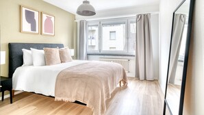 A modern bedroom with a large bed, a hanging light fixture, a window with curtains, and a mirror. - Sleek Iselin 2 Room w/ Storage, 1min to Local Market, by Blueground (Basel)