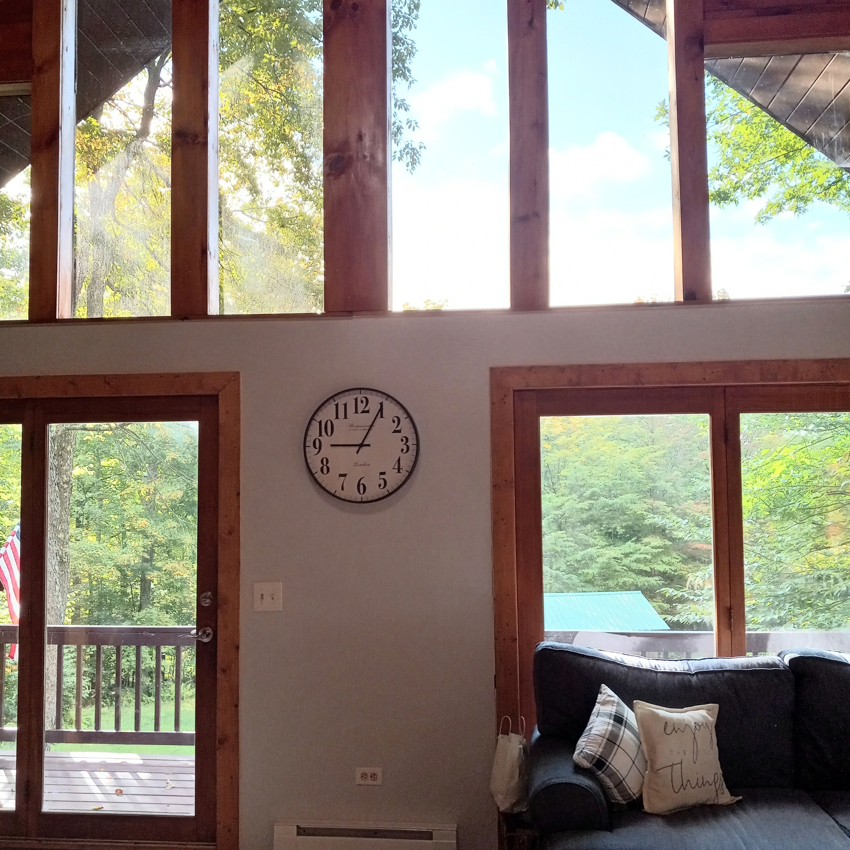 A room with a large wall clock, wooden beams, and a view of trees through large windows.