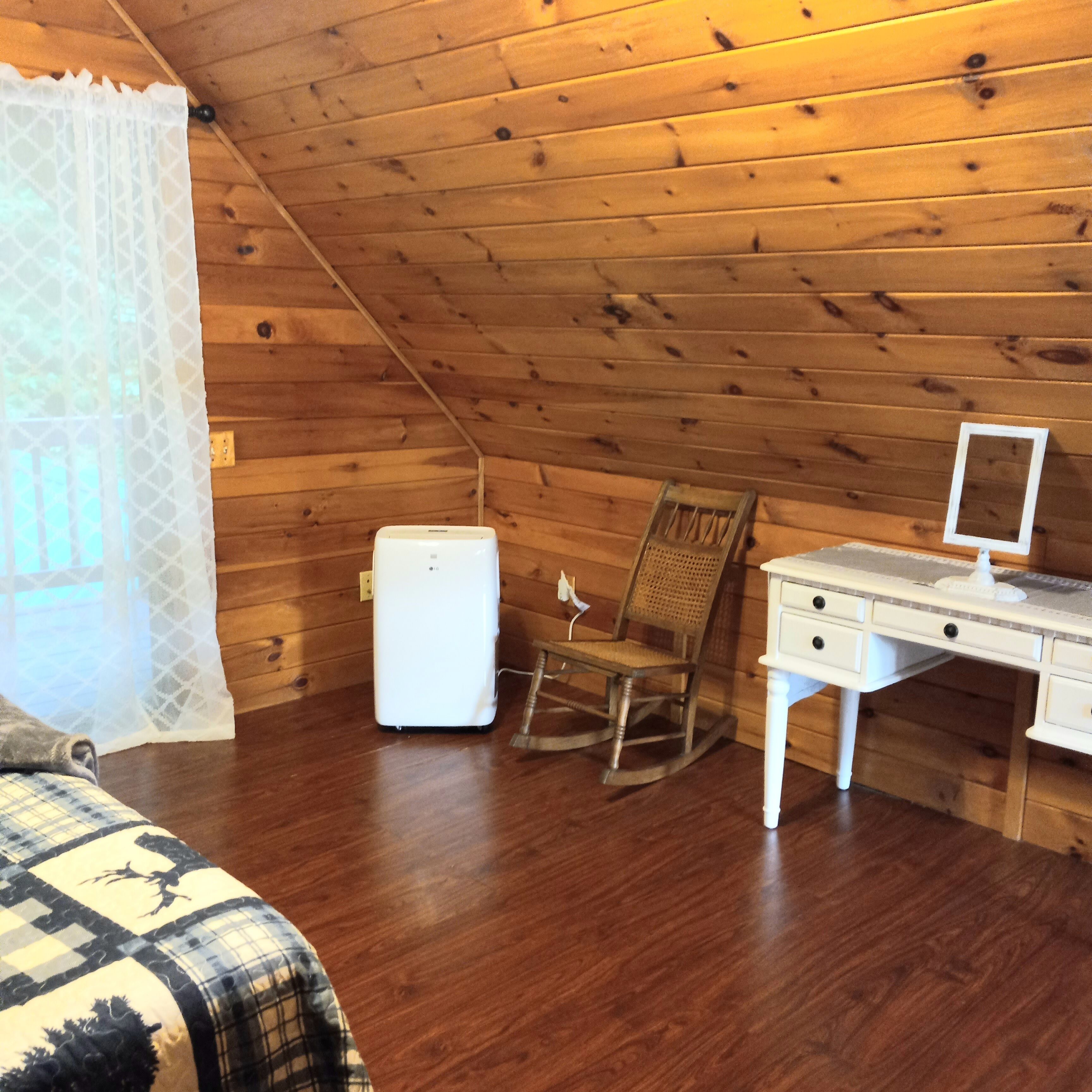 A cozy attic room with wooden walls, a white desk, a rocking chair, and a small white heater.