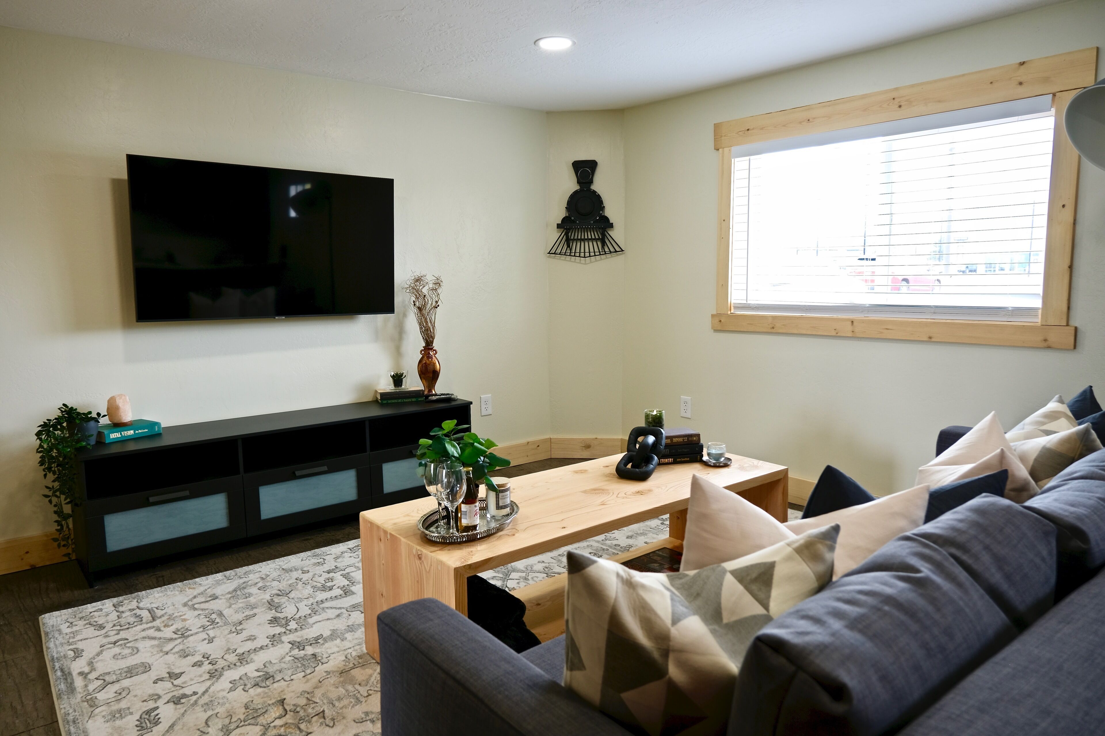 A living room with a flat-screen TV, wooden coffee table, grey sofa, and a window with blinds.