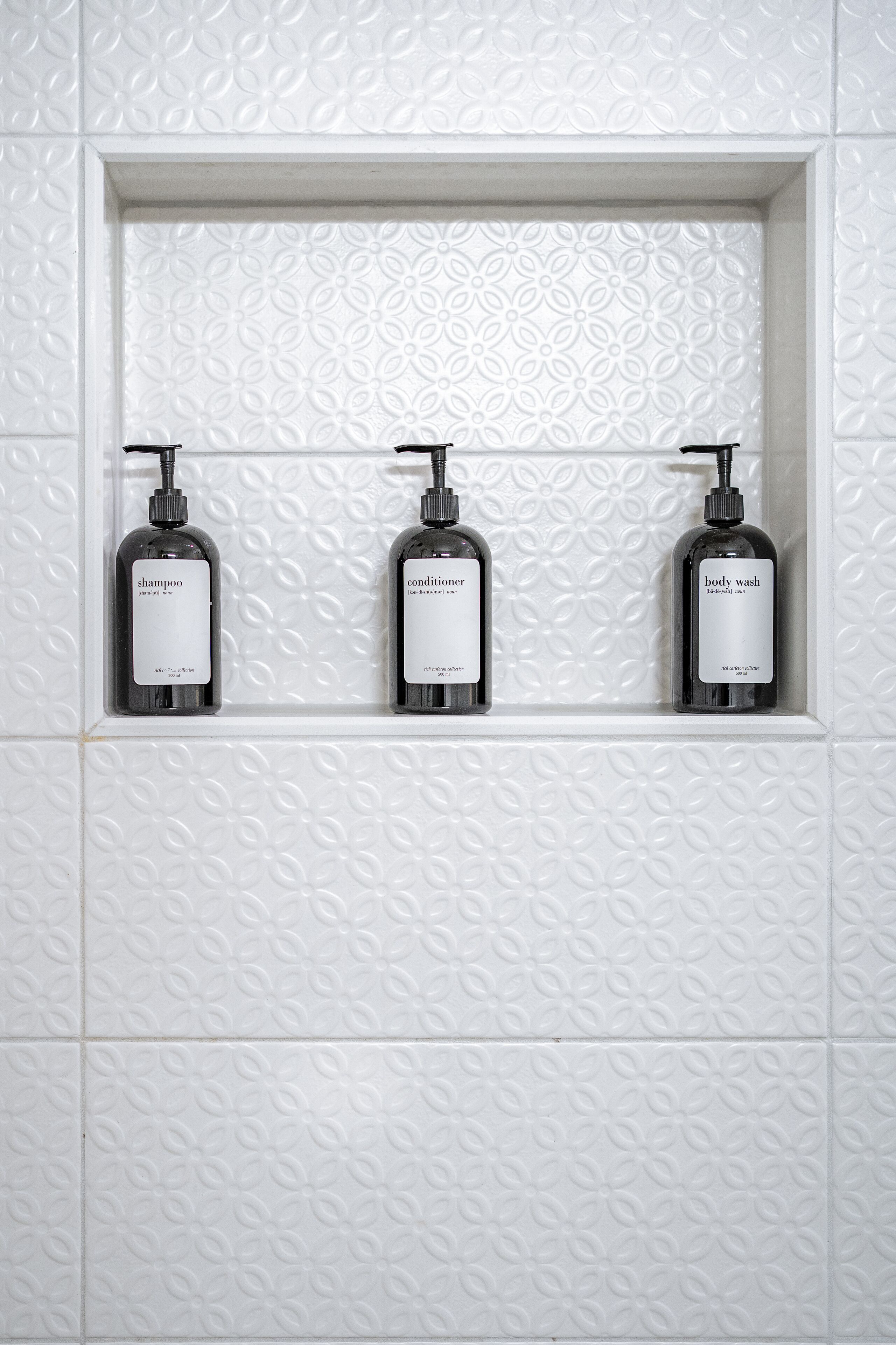 A bathroom with three bottles of shampoo, conditioner, and body wash on a white tiled wall.