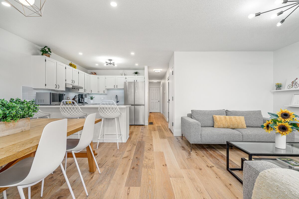 Eine moderne Küche mit weißen Schränken, ein Essbereich mit Holztisch und weißen Stühlen sowie eine Wohnbereich mit grauem Sofa und Couchtisch.