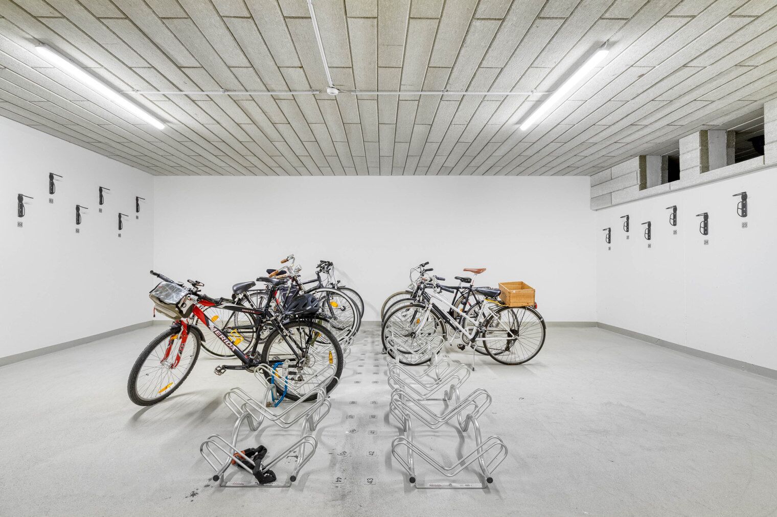 A spacious indoor area with multiple bicycles parked against the walls, a concrete floor, and a ceiling with linear lighting fixtures.