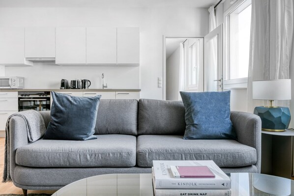 A modern living room with a grey sofa, two blue cushions, a coffee table with books, a kitchen in the background, and a window with curtains. - Bright Mariahilf 2BR w/ Elevator, near Mariahilfer Straße, by Blueground (Vienna)
