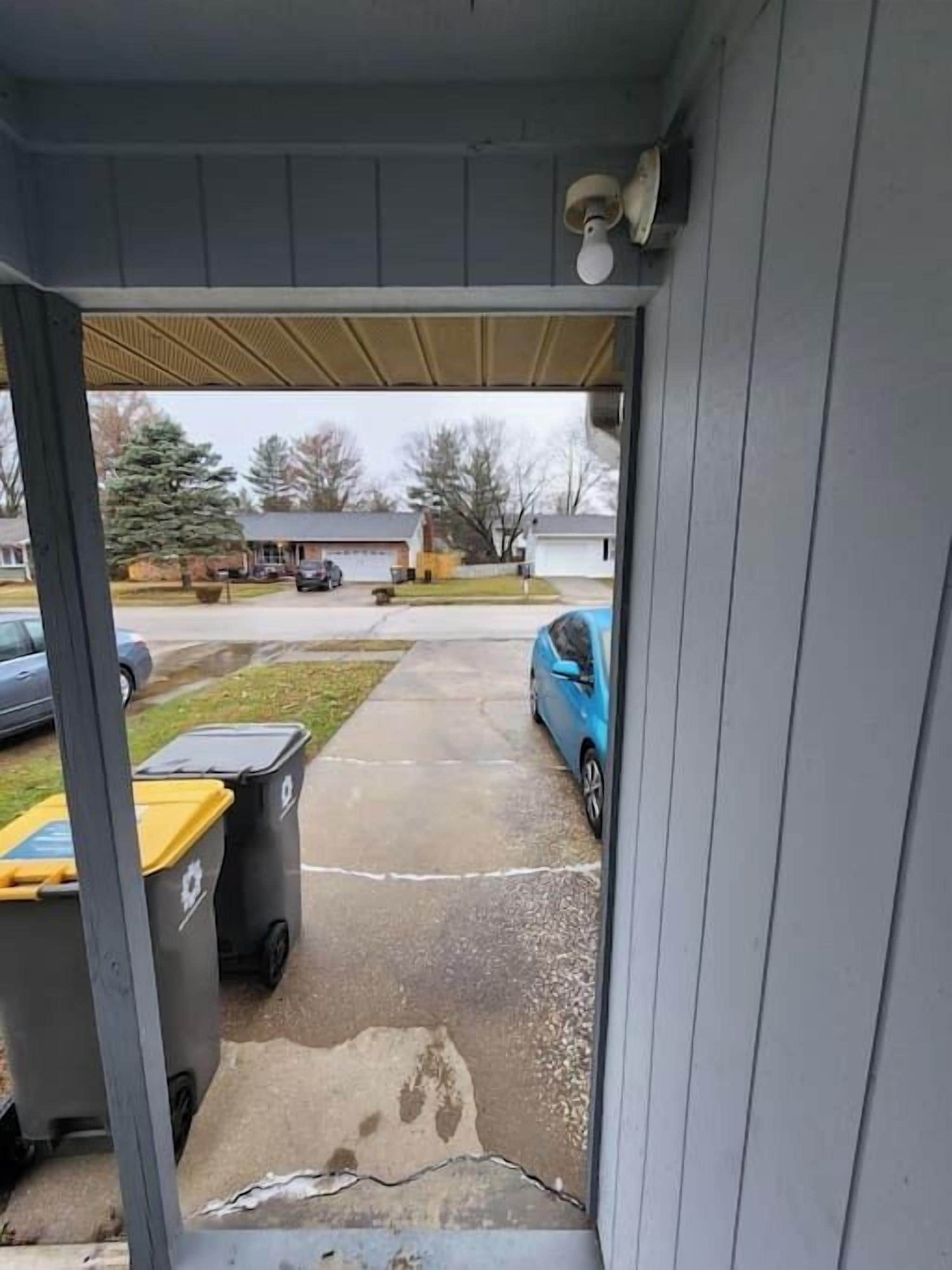 Uitzicht door een deuropening op een parkeerplaats met twee vuilnisbakken, een blauwe auto en een gebouw op de achtergrond.