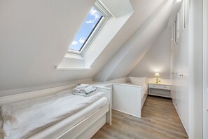 A small, white, sloped-roof bedroom with a skylight, a bed, a bedside table, and a nightstand.