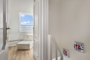 Un couloir avec une porte blanche donnant sur une pièce dotée d’une fenêtre, d’une chaise blanche et d’œuvres d’art accrochées au mur.