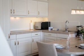 A modern kitchen with white cabinetry, a microwave, a coffee maker, and a dining area with a table set for two. - C-haus Cordula, App. 41 - Ferienwohnung mit Drei Zimmern und ca. 65 m² Wohnfläche. Für bis zu 4 (Wenningstedt-Braderup (Sylt))