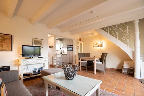 Un salon comprenant un téléviseur, un canapé et un coin repas avec une table et des chaises. Au fond, on aperçoit une cuisine équipée de placards et d’un four à micro-ondes.