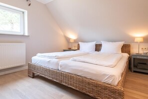 Une chambre avec un lit double, un plafond en pente, deux tables de chevet avec des lampes, un fauteuil en osier et une vue sur les arbres par la fenêtre.