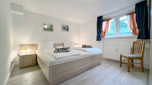 Une chambre à coucher avec un lit, une table de chevet, une lampe, une chaise et une fenêtre avec des rideaux.