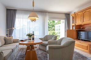 Un salon moderne avec une table ronde en bois, deux fauteuils, un canapé, un téléviseur et un coin cuisine doté de meubles en bois.