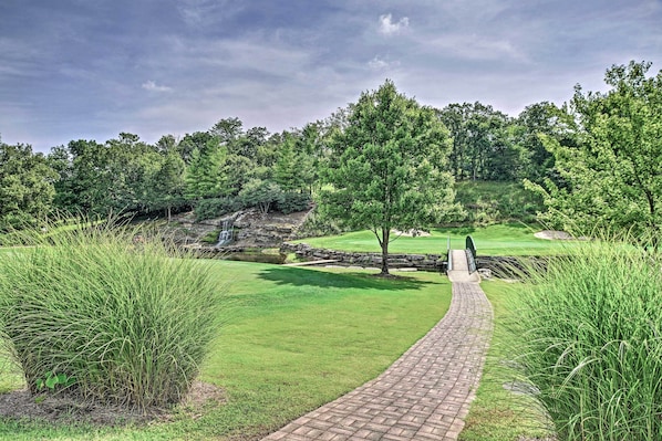 A paved pathway leading to a golf course with a pond and trees.