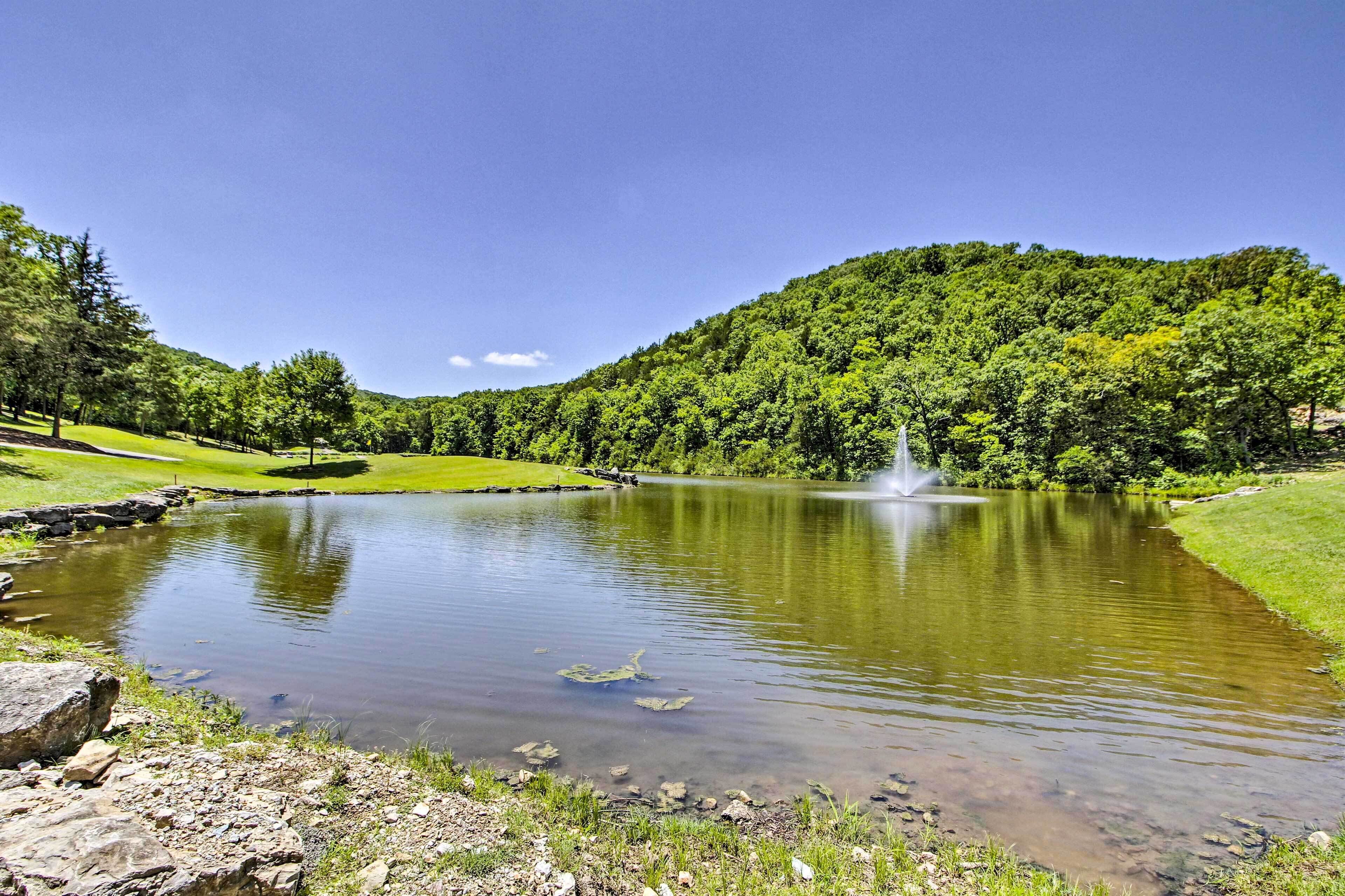 A serene lake with a fountain, surrounded by lush greenery and a clear sky.