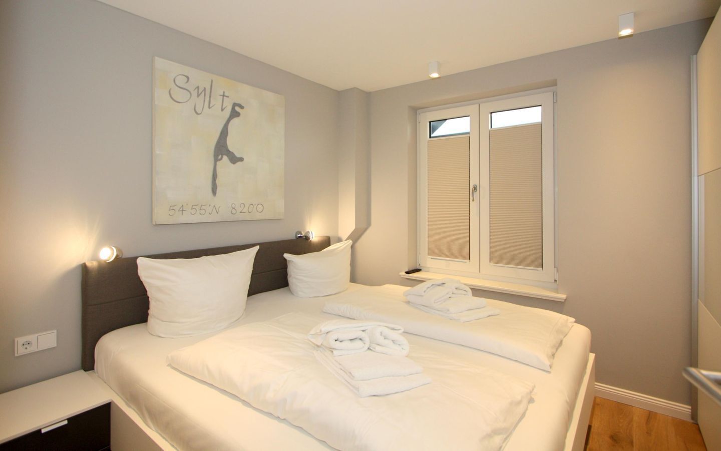 A hotel room with a large bed, two pillows, a bedside table, a nightstand, a window with blinds, and a wall-mounted artwork.