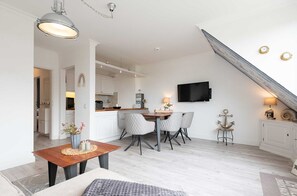 Una cocina moderna con armarios blancos, una zona de comedor con mesa y sillas de madera, y un televisor montado en la pared.
