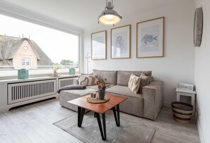 Una sala de estar acogedora con un sofá, una mesa de centro y un radiador. Hay cuadros enmarcados en la pared y una ventana con vista a un edificio.