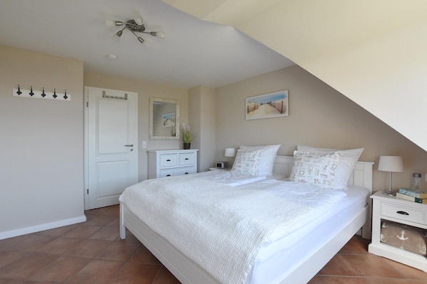 A bedroom with a large bed, a bedside table, a nightstand, a ceiling fan, a door, and a wall-mounted shelf.