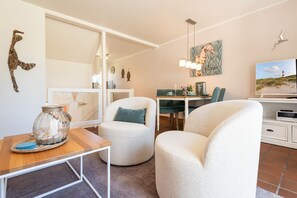 A modern living room with a sofa, a dining table, a TV, and a decorative wall piece. - Sylterpferdchen, Landhaus Köövenhüs - Gemütliche Ferienwohnung mit 3 Zimmern auf ca. 73 m², für max (Wenningstedt-Braderup (Sylt))
