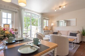 A well-lit living room with a dining area, large windows, a sofa, and a coffee table. - Sylterpferdchen, Landhaus Köövenhüs - Gemütliche Ferienwohnung mit 3 Zimmern auf ca. 73 m², für max (Wenningstedt-Braderup (Sylt))