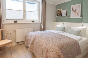 A bedroom with a bed, a chair, a radiator, and a window with blinds. - LH Haus Smer, App. 6 - Modern 3-room vacation apartment with approx. 78 m² living space, (Wenningstedt-Braderup (Sylt))