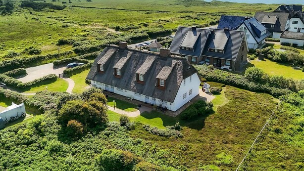 Un ambiente rurale con una grande casa bianca con tetto di paglia, circondata da vegetazione lussureggiante e una vista aperta sul mare.