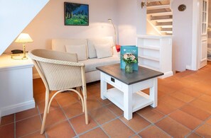 Una habitación acogedora con un sofá blanco, un sillón de mimbre, una mesita con un jarrón de flores y una escalera al fondo.