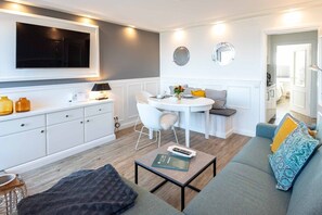 Una sala de estar moderna con un sofá gris, una mesa de centro, un comedor con mesa y sillas blancas, y una cocina con armarios blancos y un televisor montado en la pared.
