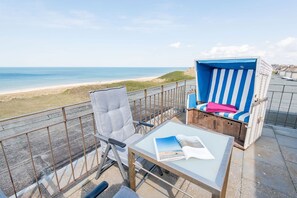 Un balcón con vista a la playa, una mesa con un libro y dos sillas de playa.