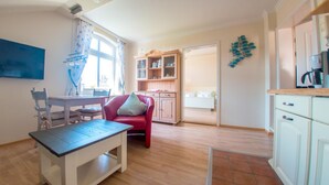 Un espacio habitable compacto con comedor, una silla roja, una cocina con armarios blancos y una puerta visible que da a otra habitación.