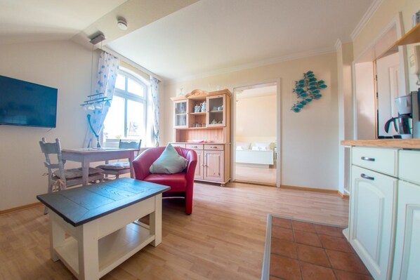 Un espace de vie compact comprenant un coin repas, une chaise rouge, une cuisine avec des meubles blancs et une porte visible donnant sur une autre pièce.