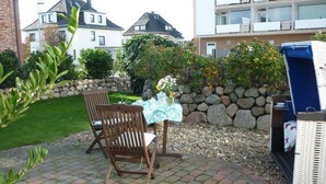 Un patio con dos sillas de madera, una mesa con mantel blanco y un pequeño jardín con muro de piedra y vegetación.