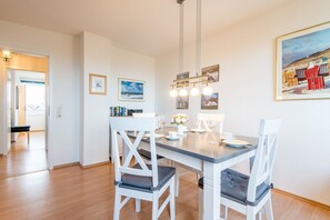 Uma sala de jantar com uma mesa de madeira, cadeiras brancas e um lustre. Há quadros nas paredes e uma pintura sobre a mesa.