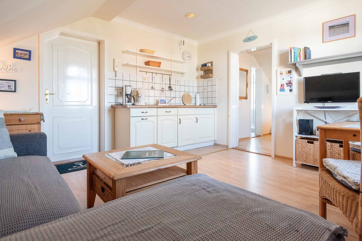 A cozy living room with a sofa, a coffee table, and a TV. There is a kitchen area with white cabinets and a tiled backsplash.