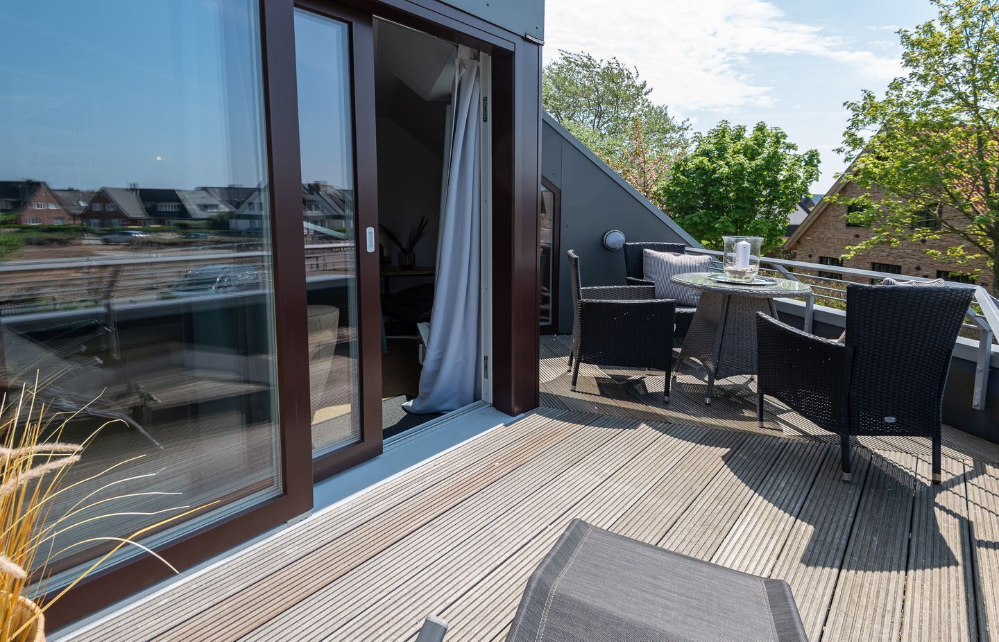 Een modern terras buiten met een glazen deur die naar een kamer leidt, gemeubileerd met rieten stoelen en een tafel.