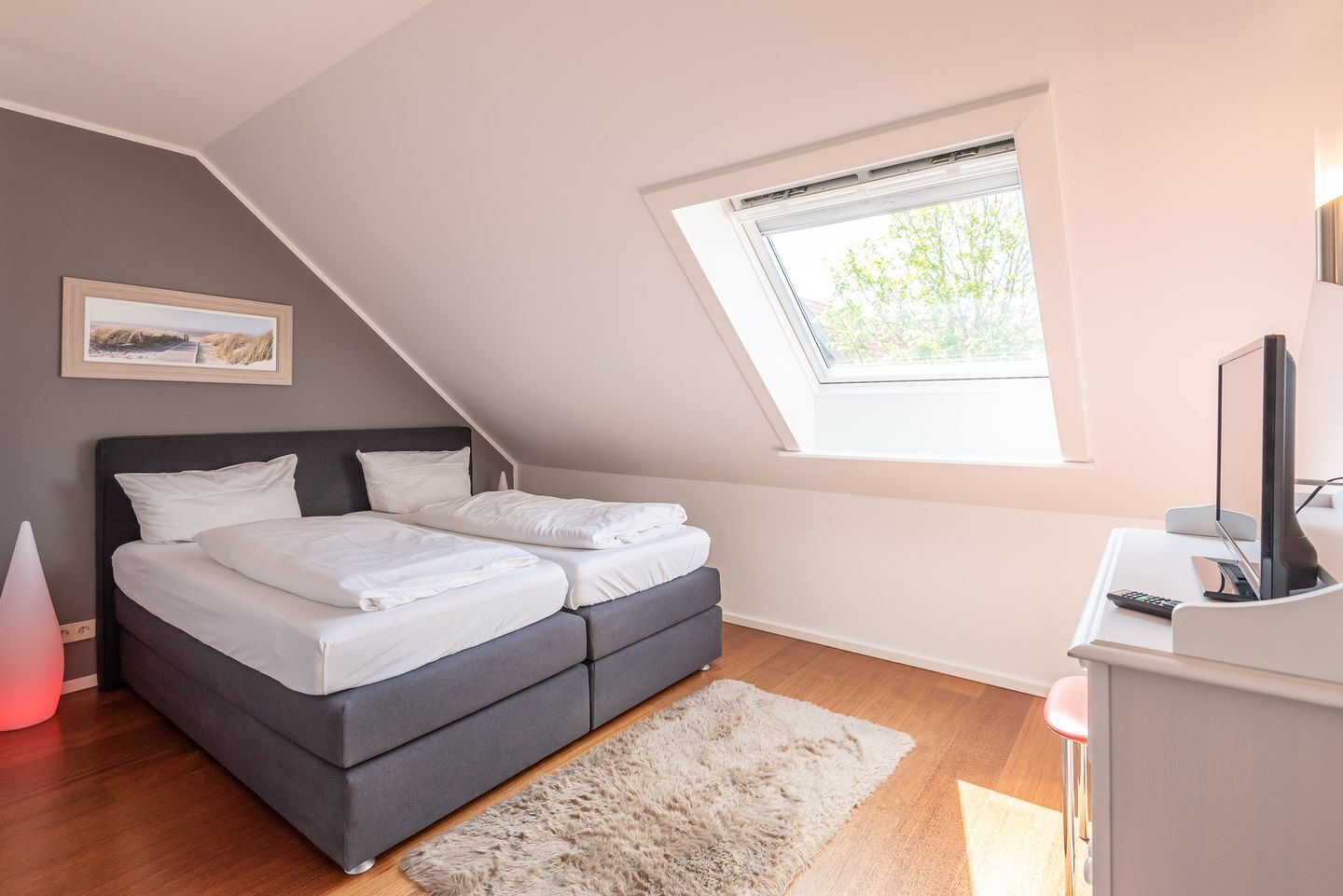 A bedroom with a bed, a desk with a computer, a window, and a painting on the wall.