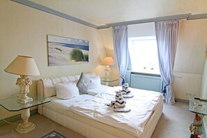 A bedroom with a large bed, two bedside lamps, a painting of a beach, and a window with curtains.