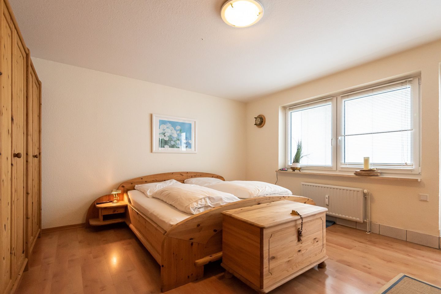 Un dormitorio con una cama de madera, una mesita de noche, un aparador, un cuadro en la pared y una ventana con persianas.