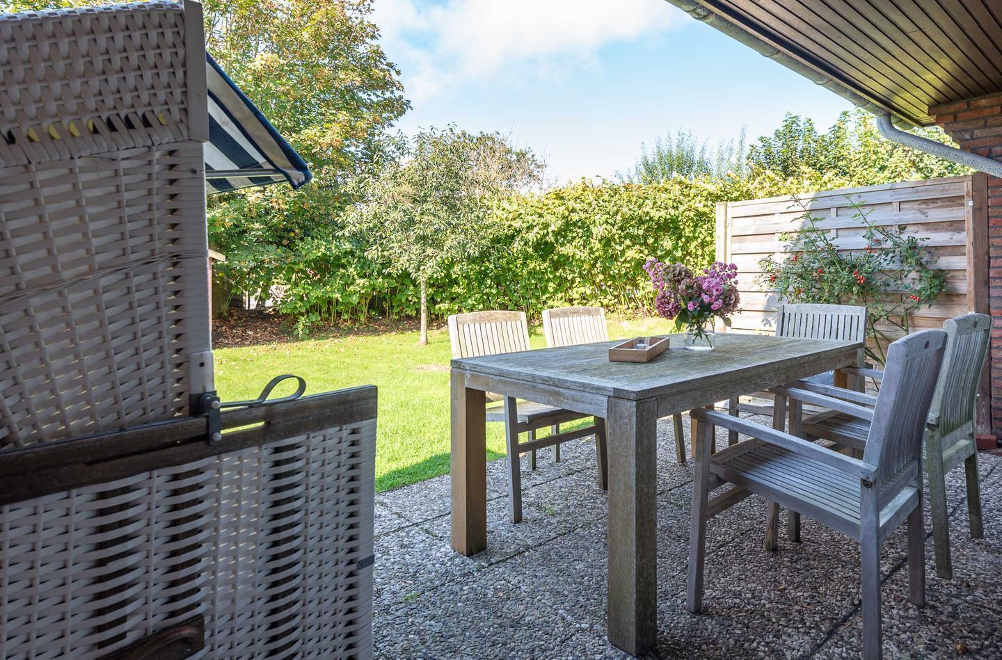 An outdoor patio with a wooden table and chairs, surrounded by greenery and a wooden fence.