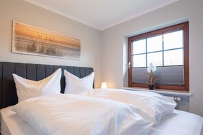 Un lit bien fait, avec du linge de lit blanc et des oreillers, une tête de lit rembourrée de couleur sombre, une grande fenêtre avec des stores et une œuvre d’art accrochée au mur représentant un coucher de soleil sur un champ.