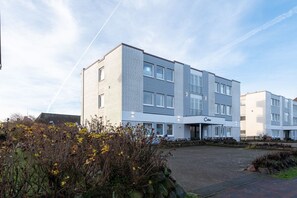 Ett modernt hotell med flera våningar, ett platt tak, stora fönster och ett välskött utomhusområde med buskar och en asfalterad uppfart.
