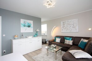 Una sala de estar moderna con un sofá de cuero marrón, una mesa de centro de vidrio y un mueble blanco. Hay un cuadro en la pared y una lámpara en la mesita.