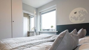 Une chambre à coucher avec un lit, des oreillers, une table de chevet, une fenêtre et un mur décoré d’une fresque représentant la lune et des papillons.