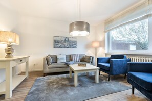 Un soggiorno moderno con un divano grigio, una poltrona blu, un tavolino da caffè bianco e un tappeto grigio. Appeso al muro c'è un quadro e una finestra con vista sugli alberi.