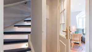 Una escalera con escalones oscuros que conduce a una habitación con una puerta blanca, una silla de mimbre y una mesita con una lámpara.
