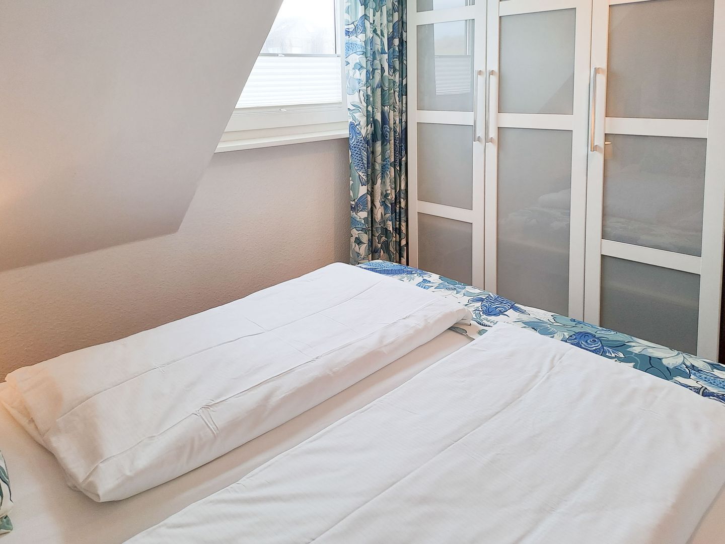 Ein kleines Zimmer mit einem Einzelbett, einem Fenster, einem Vorhang und einer weißen Tür mit satinierten Glasscheiben.
