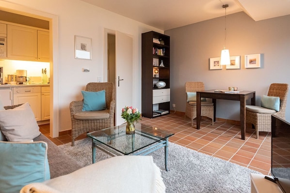 A living room with a glass coffee table, wicker furniture, a bookshelf, and a dining area with a table and chairs. - Syltdomizil Tade, App.1 - Helle 2-zimmerwohnung im Dachgeschoss mit ca. 40 m² Wohnfläche, für bis zu (Wenningstedt-Braderup (Sylt))
