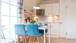 A modern kitchen with white cabinets, a stainless steel oven, and a dining area with blue chairs and a white table. - SyltStern Haus Hasenkuhle - Bright 3-room apartment with approx. 55m², for up to 4 persons. (Wenningstedt-Braderup (Sylt))