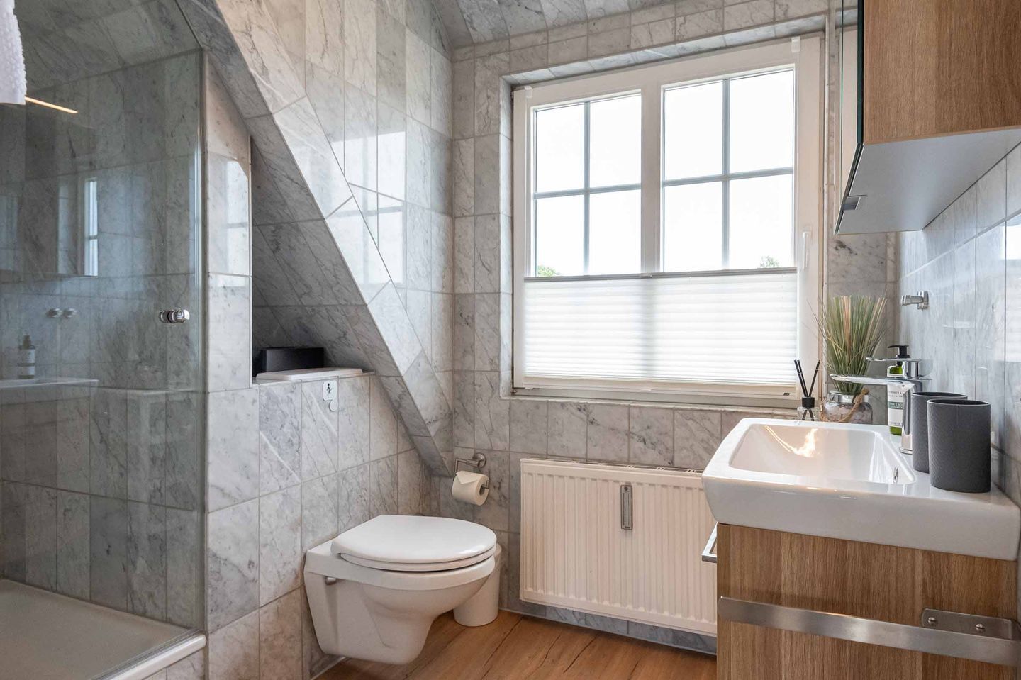 A modern bathroom with a marble floor, a white toilet, a sink with a wooden countertop, and a window with blinds.