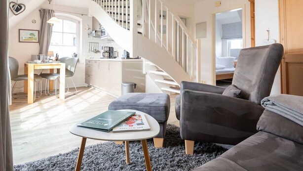 Ein modernes Wohnzimmer mit einem grauen Sofa, einem Couchtisch mit einem Buch, einer Treppe und einer Küche im Hintergrund.